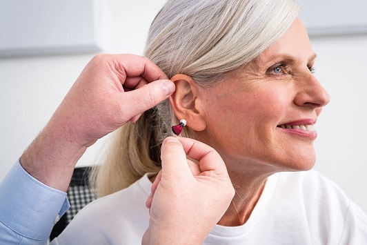 Ältere Frau bekommt ein Hörgerät ins Ohr eingesetzt und lächelt zufrieden.